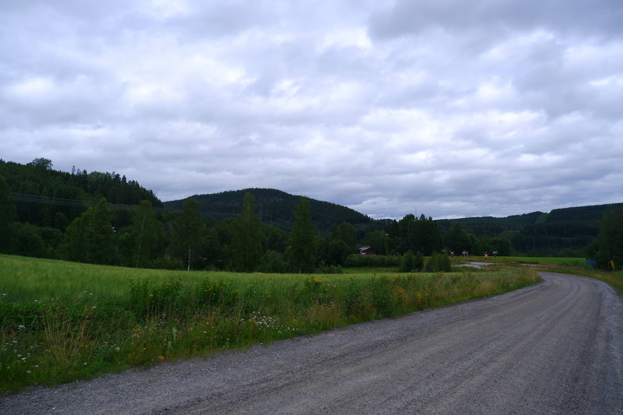 Gravel road with a railway crossing in the distance. Cloudy weather with some large hills.