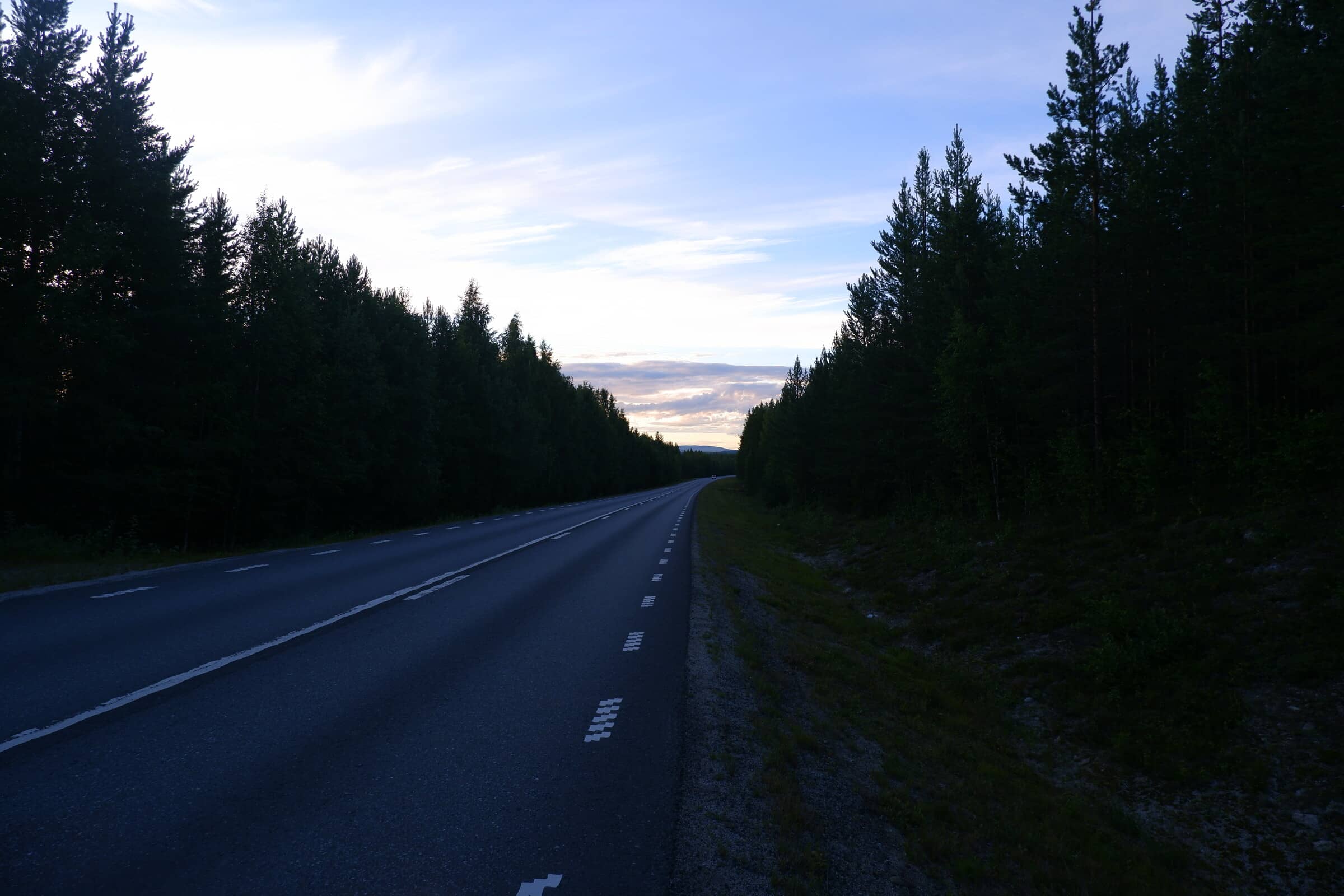 A long stretch of straight road with dense forest on the sides. The sun is coming down.